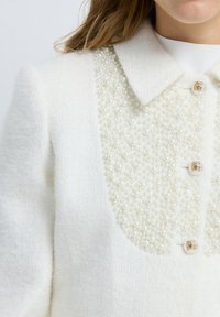 Chaqueta blanca texturizada con un panel frontal adornado con perlas, cuello clásico y detalle de botones dorados. Tejido suave y lujoso.