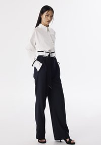 Chemise blanche à boutons avec col, associ ée à un pantalon large bleu marine foncé muni d'une taille ceinturée noire. Des sandales à talons noires complètent le look.