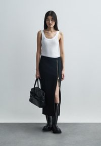 Jupe noire à fente haute avec détail de fermeture éclair dorée, associée à un débardeur blanc et des bottes noires jusqu'aux genoux. Le mannequin porte un sac à main noir.