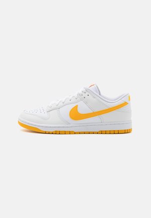 Niski biały sneaker z pomarańczową podeszwą i logo swoosh, perforowany czubek oraz białe sznurówki na jednolitym tle.