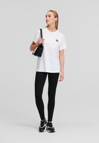 Witte katoenen T-shirt met een zwart grafisch ontwerp op de borst, gecombineerd met zwarte leggings en zwarte sneakers. Het model houdt een kleine zwarte handtas vast.