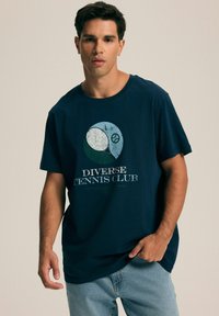 Granatowa bawełniana koszulka z okrągłym dekoltem, z graficznym wzorem przedstawiającym elementy tenisa oraz napisem "Diverse Tennis Club" w postarzanym białym druku.