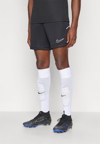 Μαύρη σορτς Nike με λευκό λογότυπο, λευκές κάλτσες με μαύρες λεπτομέρειες, και μαύρα και μπλε ποδοσφαιρικά παπούτσια με υφή και καρφιά.