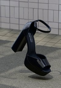 Μαύρα πέδιλα από πατέντα δέρμα με ψηλό τακούνι και πλατφόρμα, ανοιχτή μύτη, λουράκι στον αστράγαλο και χοντρό τακούνι. Λευκή, γυαλιστερή επιφάνεια.