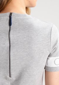 Grå kortärmad T-shirt med dragkedja baktill, med en mjuk textur och minimalistisk design. Vit detalj på ärmen.