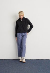 Person with short blonde hair wearing a black jacket, light blue pants, white socks, and white sneakers standing against a white wall.
