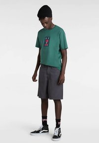 Groen katoenen T-shirt met een grafische print op de borst, gecombineerd met grijze shorts. Zwarte beanie, zwarte sokken met roze letters en zwarte sneakers.