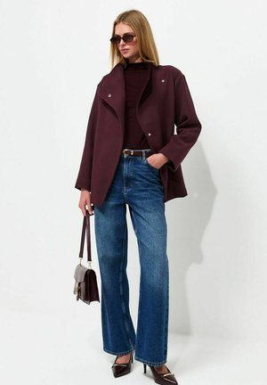 Mujer con abrigo rojo oscuro, parte superior oscura, vaqueros azules de pierna ancha, gafas de sol y tacones altos, sosteniendo un bolso oscuro en una mano.