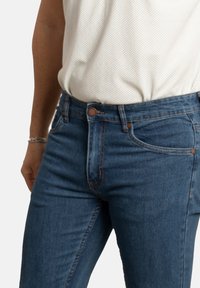 Jeans de denim azul oscuro para hombre con un corte slim, que cuentan con un diseño clásico de cinco bolsillos, costuras en color marrón y cierre delantero con botón.