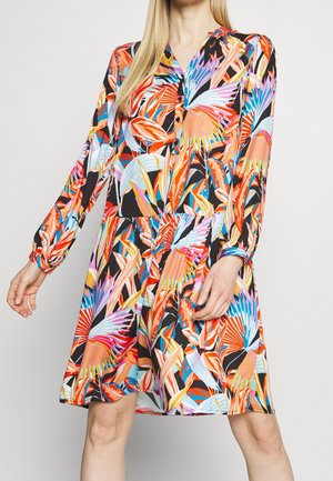 Femme portant une robe colorée à manches longues avec un motif de feuilles abstrait et vibrant en orange, bleu, rose et noir.