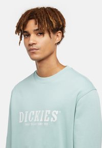 Sudadera azul claro con cuello redondo, que presenta un logotipo bordado en blanco "DICKIES" y el texto "CONSTRUIDA PARA DURAR DESDE 1922" en el pecho.