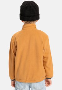 Oranžová fleecová bunda s vysokým límcem, s černým lemem na manžetách, nošená s světle modrými džínami a černou pletenou čepicí.