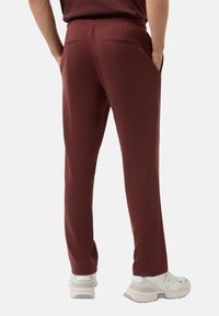 Bordeauxkleurige broek met een gladde textuur, een elastische tailleband en twee achterzakken. Gecombineerd met lichtgekleurde sneakers.