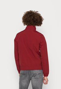 Rote leichte Jacke mit Stehkragen, elastischen Bündchen und Saum; von hinten mit grauen Jeans präsentiert.