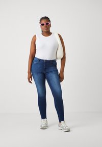 Ψηλοκάβαλες μπλε τζιν παντελόνες, εφαρμοστό σχέδιο, αντίθετη ραφή και πέντε τσέπες, συνδυασμένες με λευκή μακρυμάνικη μπλούζα και στρογγυλά γυαλιά ηλίου.