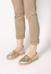 Metalen gouden espadrilles met uitgesneden bloemmotieven, tan accentriem en gevlochten jute zool, gedragen met opgerolde beige broek.