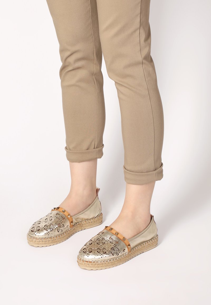 Metalen gouden espadrilles met uitgesneden bloemmotieven, tan accentriem en gevlochten jute zool, gedragen met opgerolde beige broek.