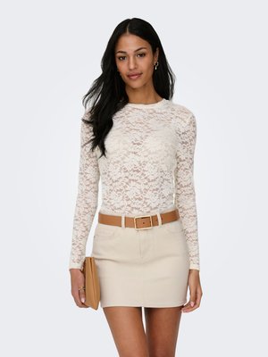 Kobieta o długich czarnych włosach ubrana w białą koronkową bluzkę z długim rękawem, beżową mini spódniczkę z brązowym paskiem, złote kolczyki koła, trzyma beżową kopertówkę.