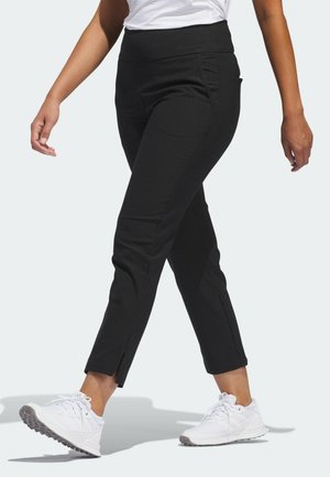 Mujer caminando con pantalones ajustados negros con aberturas en los tobillos, zapatillas blancas y una camiseta blanca metida por dentro.