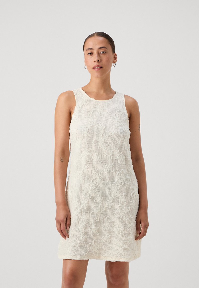 Robe blanche sans manches en tissu texturé, ornée de broderies florales et d'une coupe décontractée. Surface lisse et douce avec un design léger.