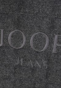 Szara filcowa tkanina ma wytłoczone logo "JOOP" nad "JEANS", z teksturowaną powierzchnią i przeszyciami.