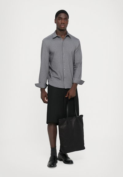 Uomo che indossa una camicia grigia con bottoni e pantaloni corti neri fino al ginocchio, mentre tiene una grande borsa a tracolla nera, in piedi contro uno sfondo bianco.