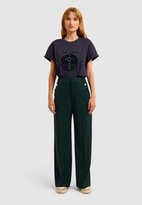 T-shirt navy avec impression graphique, manches courtes. Pantalons larges verts avec boutons sur le devant et poches latérales. Chaussures beiges unies.