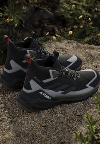 Μαύρα και γκρι ορειβατικά παπούτσια Adidas Terrex Gore-Tex τοποθετημένα σε χαλίκι με πράσινο φύλλωμα στο φόντο.
