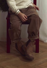 Brūni ādas potītes ar elastīgām sānu paneļiem, valkātas ar gaiši brūnām kordūra biksēm, sēžot uz sarkanā krēsla ar koka grīdu.