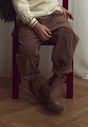 Bottines en cuir marron avec panneaux élastiques sur les côtés, portées avec un pantalon en velours côtelé marron clair, assises sur une chaise rouge avec un sol en bois.