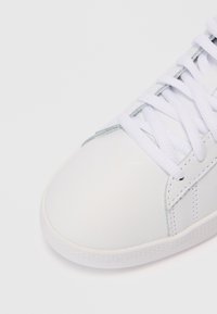 Zapatilla blanca que presenta un material sintético liso, punta redonda y cordones planos. Los acentos laterales incluyen líneas cosidas para un detalle adicional.