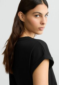 T-shirt nero con una vestibilità rilassata e maniche arrotolate, caratterizzato da una texture morbida. I lunghi capelli mossi del modello scendono su una spalla.