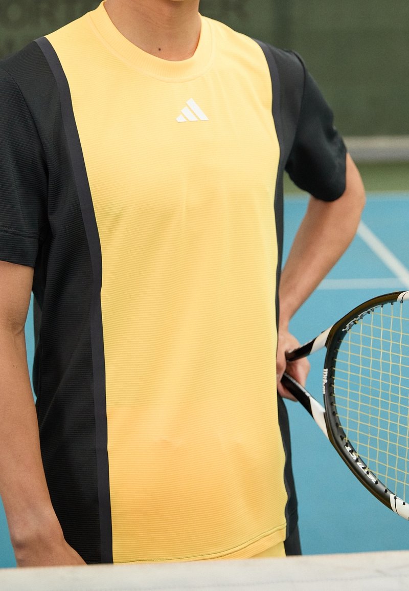 Camiseta de tenis de manga corta amarilla y negra con una textura acanalada, que presenta una costura central y el logo de Adidas en el pecho.