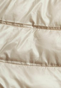 Tejido de chaqueta acolchada color beige claro con una textura suave y brillante. Presenta un acolchado horizontal y costuras visibles a lo largo de las uniones.