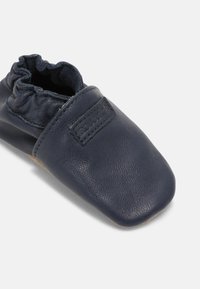 Scarpa per neonati in pelle blu navy con una finitura morbida e testurizzata e colletto elastico arricciato. Include una piccola etichetta con il marchio sulla parte superiore.