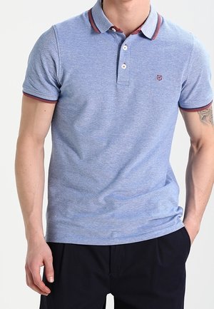 Homme portant un polo bleu clair à manches courtes avec une bordure rouge foncé au col et aux manches, associé à un pantalon noir.