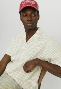 Camisa de manga corta, con cuello, en un beige claro con un patrón de cuadros. Modelo con una gorra roja, sentado en una silla con respaldo de madera.
