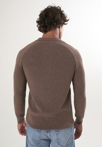 Pull en maille côtelée marron avec des manches longues et un col rond, présentant une silhouette ajustée et un tissu texturé sur fond clair.
