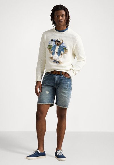 Fehér kötött pulóver, színes maci mintával, világoskék farmer rövidnadrággal és kék sportcipővel, fehér fűzővel párosítva.