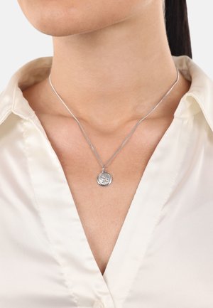 Silberne Anhängerkette mit einem runden Charm mit einem geprägten Design, hängend an einer feinen Kette, getragen über einem cremefarbenen Hemd.