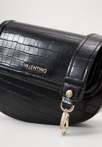 Svart handväska i konstläder med krokodilprägling, rund form, guldaccessoarer, nyckelringklämma och viklock. Innehar logotypdetalj.