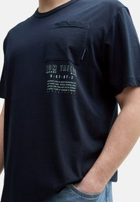 Granatowa bawełniana koszulka z krótkim rękawem, z kieszonką na piersi i nadrukowanym napisem. Charakteryzuje się luźnym krojem i miękką teksturą.