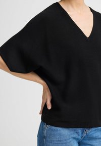 Haut noir à col V avec des manches larges et courtes, fabriqué dans un tissu lisse, présentant un ourlet droit, associé à un jean bleu clair.