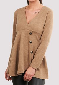 Beige strikket topp med dyp V-hals, omslagsdesign, lange ermer og tre mørke knapper på siden. Myk tekstur med en flytende silhuett.