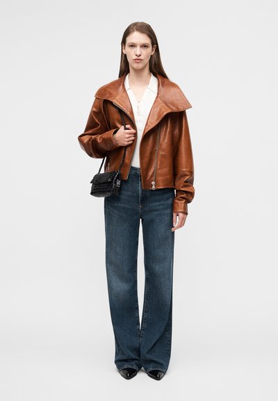 Καφέ δερμάτινο μπουφάν με φαρδιά γιακά, ανοιχτό μπροστινό φερμουάρ και μακριά μανίκια, συνδυασμένο με μπλε τζιν παντελόνι φαρδιάς γραμμής και μικρή μαύρη τσάντα.