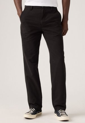 Pantalones de tela de algodón negro con un diseño recto, que presentan bolsillos laterales, una textura suave y se llevan con zapatillas negras y blancas.