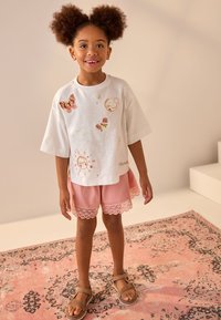 Bílé tričko s krátkým rukávem s různými vyšívanými motivy, doplněné růžovými šortkami s krajkovým lemem. Hnědé sandály na bosých nohách.
