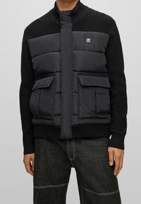 Giacca a vento nera con maniche in maglia a costine, colletto alto, due tasche anteriori e una patch con logo sul petto. Finitura opaca e texture imbottita.