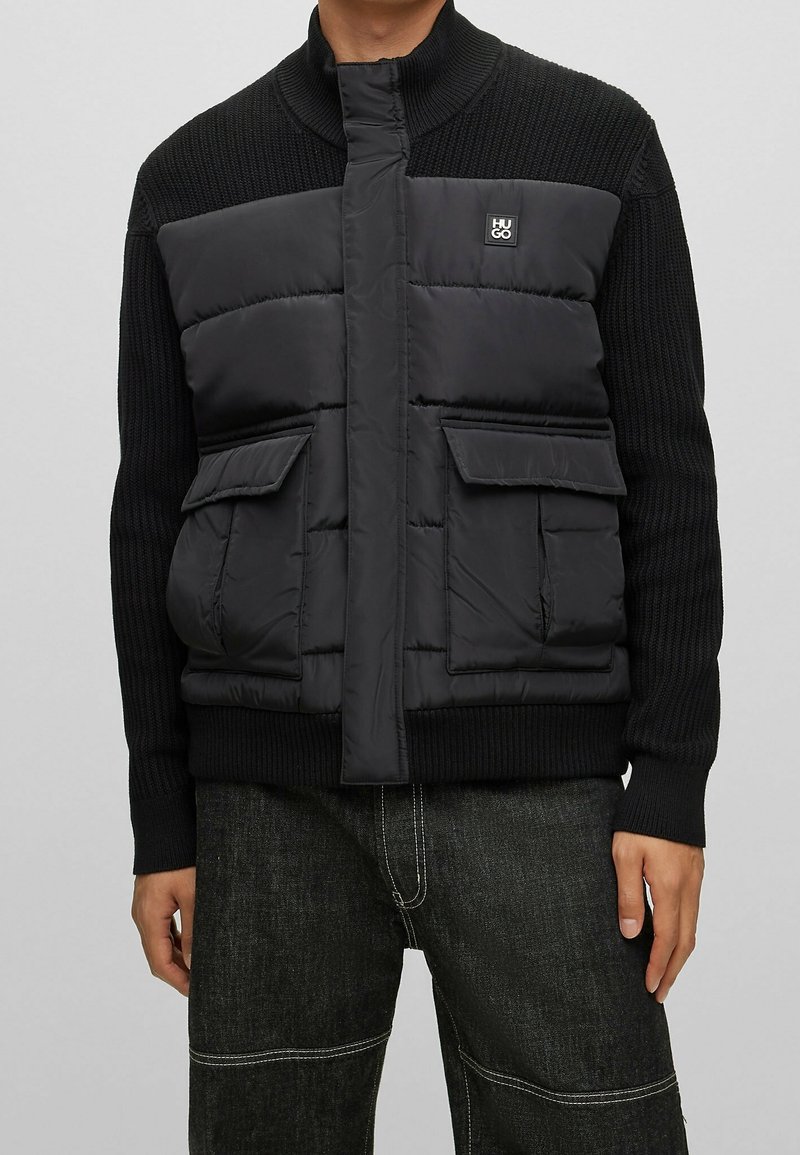 Giacca a vento nera con maniche in maglia a costine, colletto alto, due tasche anteriori e una patch con logo sul petto. Finitura opaca e texture imbottita.