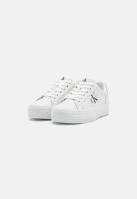 White leather sneakers with a round toe, flat laces, and a textured rubber sole. Features a logo on the side and contrasting accents.